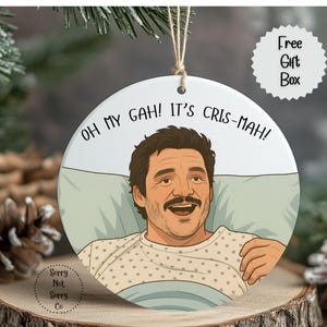 May include: A round Christmas ornament featuring a cartoon illustration of a man with a mustache in a hospital bed. The ornament has the text "OH MY GAH! IT'S CRIS-MAH!" and a "Free Gift Box" label.