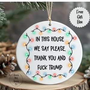 May include: A white ceramic Christmas ornament with the text "IN THIS HOUSE WE SAY PLEASE, THANK YOU AND FUCK TRUMP" in black. The ornament is decorated with colorful string lights and a small "Free Gift Box" graphic.