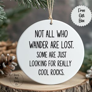 Puede incluir: Adorno redondo de cer&aacute;mica blanca con el texto "NOT ALL WHO WANDER ARE LOST. SOME ARE JUST LOOKING FOR REALLY COOL ROCKS." Colgando de una cuerda, est&aacute; sobre una superficie de madera.