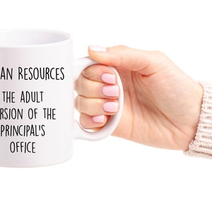 May include: White ceramic mug with the text "HUMAN RESOURCES THE ADULT VERSION OF THE PRINCIPAL'S OFFICE" in black. The mug is held by a person.