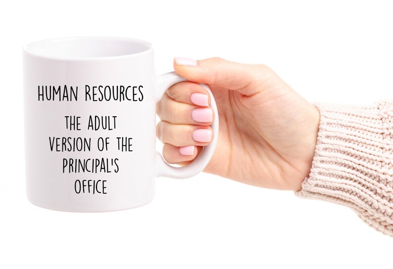 May include: White ceramic mug with the text "HUMAN RESOURCES THE ADULT VERSION OF THE PRINCIPAL'S OFFICE" in black. The mug is held by a person.