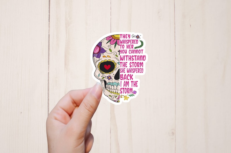 May include: A sticker with a colorful sugar skull design and the text "They whispered to her you cannot withstand the storm she whispered back I am the storm".
