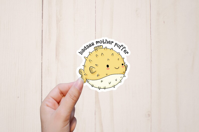 Puede incluir: Un pez globo amarillo y blanco con una cara sonriente y el texto "badass mother puffer" en negro.