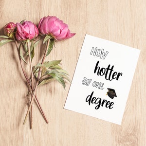 May include: A greeting card with the text "Now hotter by one degree" and a graduation cap illustration. The card is next to pink peonies with green leaves and stems, all on a light wood surface.