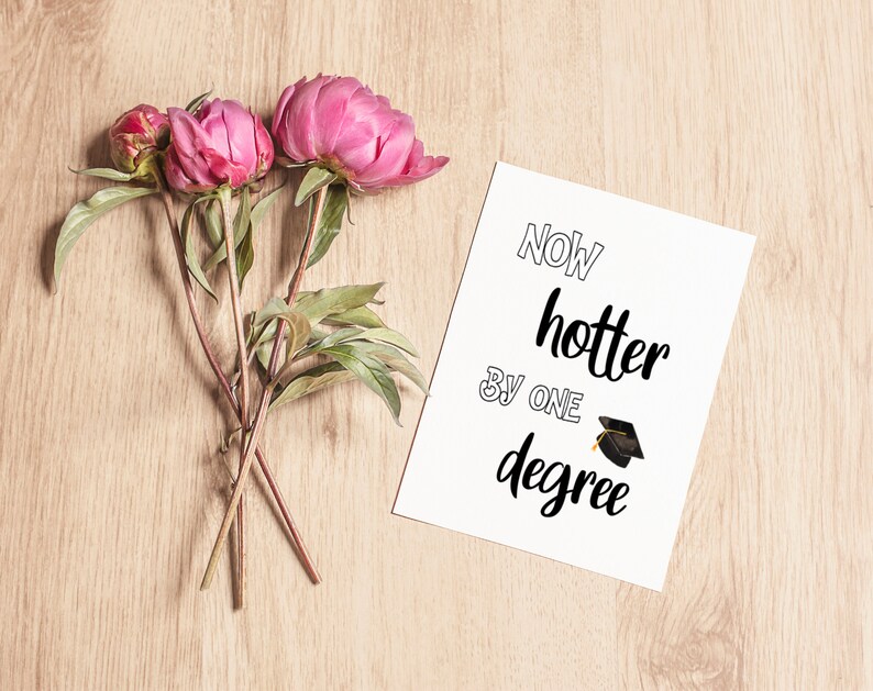May include: A greeting card with the text "Now hotter by one degree" and a graduation cap illustration. The card is next to pink peonies with green leaves and stems, all on a light wood surface.