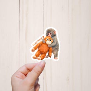 May include: A sticker featuring two cartoon monkeys. One gray monkey hugs an orange monkey. The text "TEAM PUNCH!" is printed in brown with a heart symbol. The sticker is held by a hand.