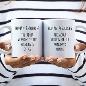 May include: Two white ceramic mugs with black text that reads "HUMAN RESOURCES THE ADULT VERSION OF THE PRINCIPAL'S OFFICE." The mugs have a curved handle and are held up by a person.