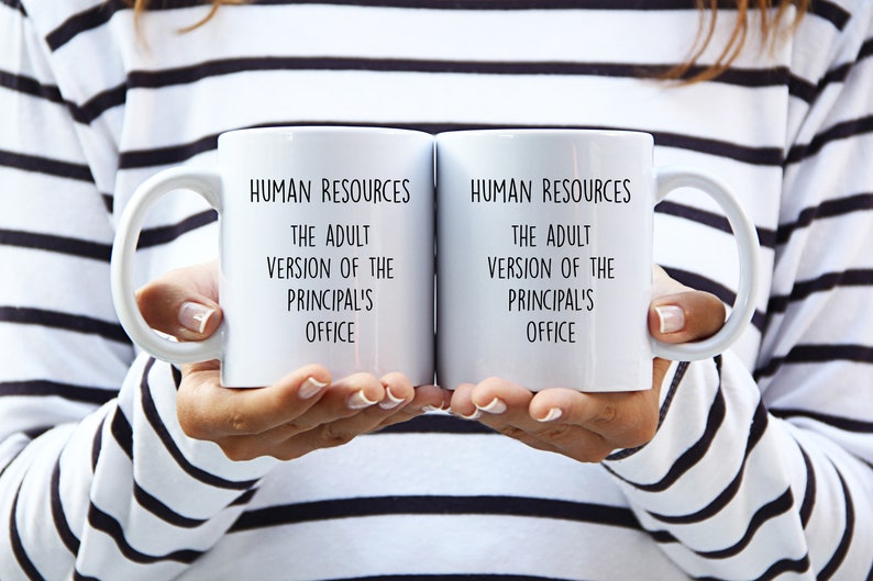 May include: Two white ceramic mugs with black text that reads "HUMAN RESOURCES THE ADULT VERSION OF THE PRINCIPAL'S OFFICE." The mugs have a curved handle and are held up by a person.