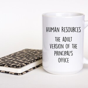 May include: A white ceramic mug with the text "HUMAN RESOURCES THE ADULT VERSION OF THE PRINCIPAL'S OFFICE" in black. A closed notebook with a leopard print cover is next to the mug.
