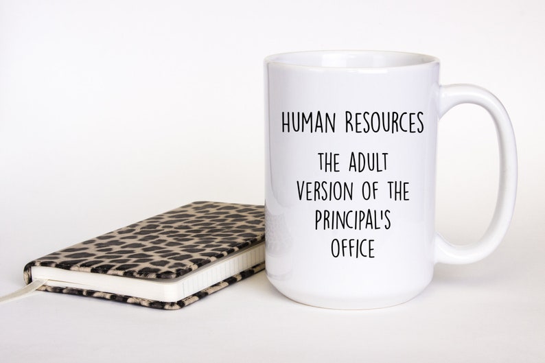 May include: A white ceramic mug with the text "HUMAN RESOURCES THE ADULT VERSION OF THE PRINCIPAL'S OFFICE" in black. A closed notebook with a leopard print cover is next to the mug.