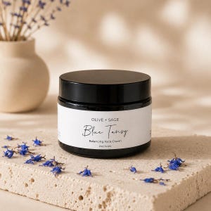 Closed jar of Olive + Sage Blue Tansy Balancing Face Cream on a warm stone surface with scattered blue flowers and a soft beige botanical background.