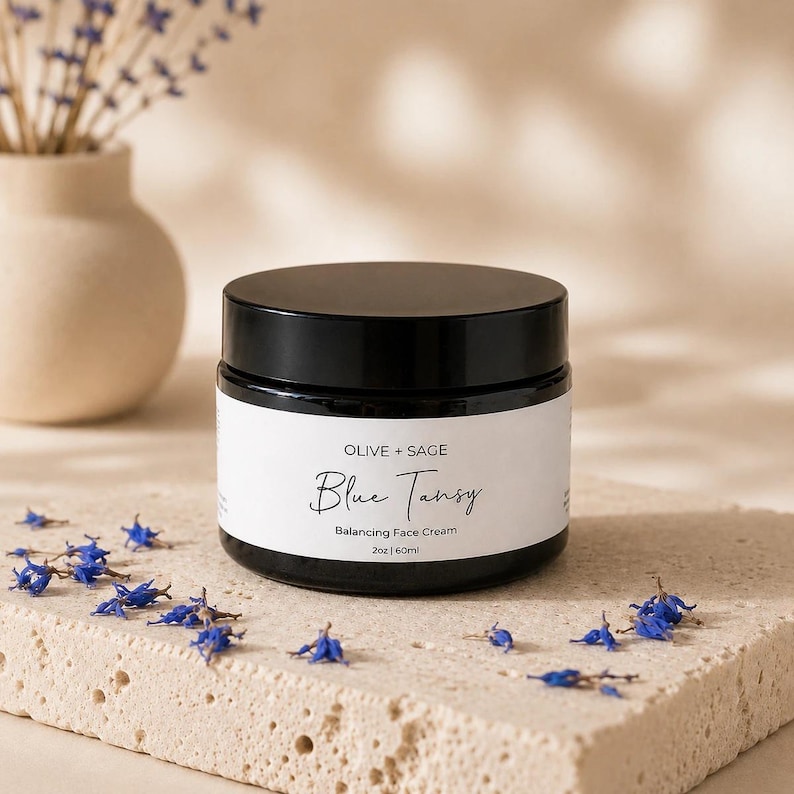 Closed jar of Olive + Sage Blue Tansy Balancing Face Cream on a warm stone surface with scattered blue flowers and a soft beige botanical background.