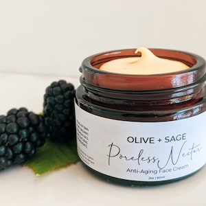 Op de afbeelding: Een bruine glazen pot met een wit etiket waarop staat "OLIVE + SAGE Poreless Nectar Anti-Aging Gezichtscrème 2oz | 60ml". De pot is gevuld met een gele crème en staat op een wit oppervlak met bramen.