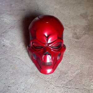 Puede incluir: Un casco rojo con forma de cráneo con detalles negros y una visera. El casco tiene un aspecto amenazante y está hecho de un material plástico duro.