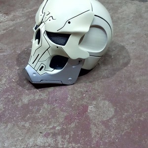 Pode incluir: Um capacete branco em forma de caveira com detalhes em preto e prata. O capacete tem uma viseira e uma superf&iacute;cie texturizada.