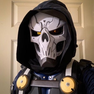 Pode incluir: Uma m&aacute;scara de caveira branca com olhos amarelos, usada sobre uma roupa com capuz preto. A m&aacute;scara tem um design detalhado com bordas afiadas e uma superf&iacute;cie texturizada. A roupa tem um capuz preto e um tecido azul escuro por baixo.