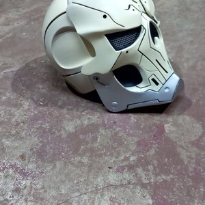 Pode incluir: Um capacete branco com um design de caveira e detalhes em preto e prata. O capacete tem uma viseira de malha e uma superf&iacute;cie texturizada.
