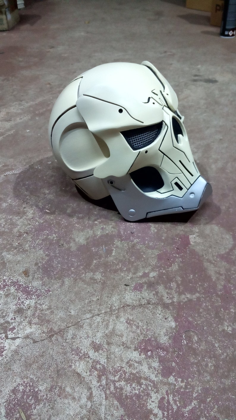 Pode incluir: Um capacete branco com um design de caveira e detalhes em preto e prata. O capacete tem uma viseira de malha e uma superf&iacute;cie texturizada.
