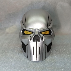 Puede incluir: Una máscara de calavera plateada con detalles negros y cuencas de los ojos de malla amarilla. La máscara tiene un acabado metálico y un diseño amenazante, adecuado para cosplay o exhibición.