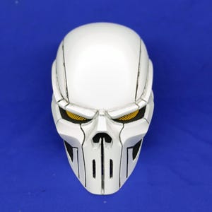 Puede incluir: Una máscara de calavera blanca con detalles en negro y cubiertas oculares de malla amarilla. La máscara tiene un diseño angular detallado con ranuras verticales para la boca. Probablemente para cosplay o disfraces.