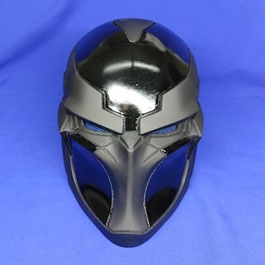 Puede incluir: Una máscara futurista negra y gris con un acabado brillante. La máscara tiene cubiertas oculares de malla azul y un diseño elegante y angular. La parte superior de la máscara es de un negro más oscuro, mientras que la parte inferior es de un gris más claro. La máscara está diseñada para cubrir toda la cara.