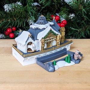 May include: A detailed Christmas-themed toy workshop with a snow-covered roof, a rocking horse, and the words "Toy Workshop." The miniature scene includes a teddy bear wearing a Santa hat and small green trees.