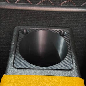 May include: A black cup holder insert with a carbon fiber pattern finish, featuring a circular opening and four corner screw holes, mounted within a black textured console housing adjacent to a bright yellow leather-textured surface.