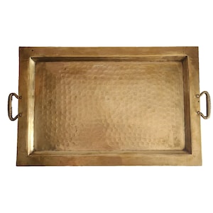 May include: A rectangular brass tray featuring a hammered texture on the base and a smooth, flat border. The tray includes two small, curved, ribbed handles attached to the sides with visible rivets, showcasing a metallic gold finish.