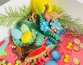 May include: A pink sensory bin filled with sand, blue and yellow plastic eggs, green and blue glass gems, and toy dinosaurs. The bin also includes green plastic plants, brown paper shreds, and colorful plastic flowers.