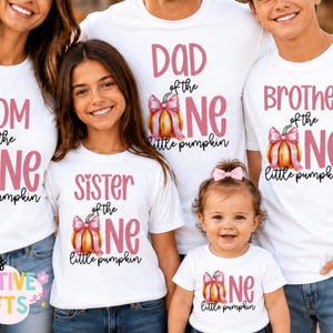 May include: A group of five white t-shirts featuring pink text and a pumpkin graphic with a pink bow. The shirts display the phrases Mom of the one little pumpkin, Dad of the one little pumpkin, Sister of the one little pumpkin, Brother of the one little pumpkin, and One little pumpkin.