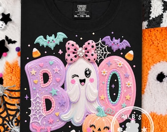 May include: A black t-shirt featuring a graphic print of the word BOO, where the letter O is replaced by a winking ghost wearing a pink polka-dot bow. The design includes pastel-colored bats, spiderwebs, stars, flowers, and a jack-o'-lantern.