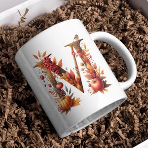 May include: White ceramic mug with a fall-themed design featuring the letter "W" surrounded by autumn leaves, berries, and a small pumpkin.