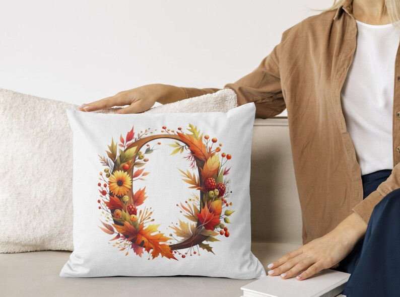 May include: White throw pillow with a fall-themed design featuring a wreath of orange, yellow, and red leaves, berries, and flowers. The wreath surrounds a brown letter "O".