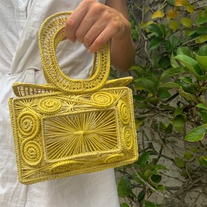 May include: A yellow woven handbag with a rectangular body and a rounded handle. The bag features a sunburst design in the center and spiral details on the sides. The bag is made of a woven material.