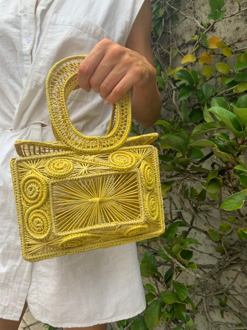 May include: A yellow woven handbag with a rectangular body and a rounded handle. The bag features a sunburst design in the center and spiral details on the sides. The bag is made of a woven material.