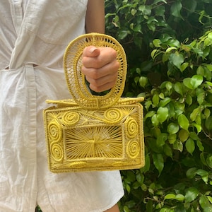 May include: A yellow woven handbag with a rectangular body and a circular handle. The bag features intricate spiral and linear patterns. The bag is held in front of a green leafy background.