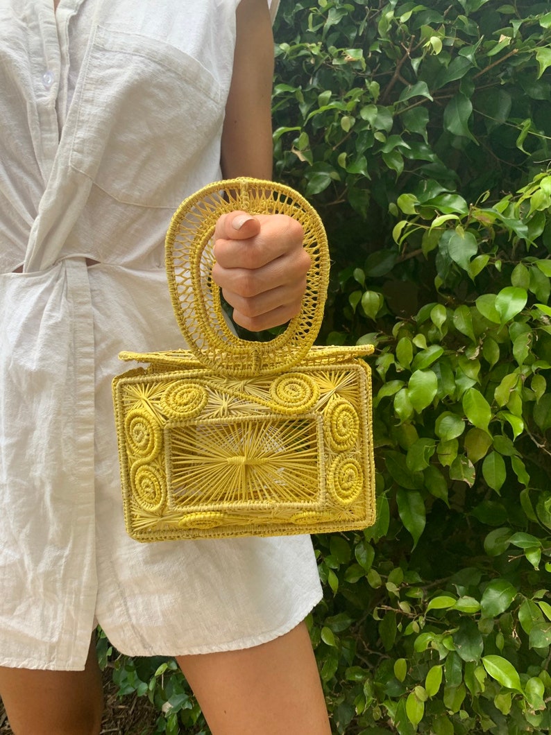 May include: A yellow woven handbag with a rectangular body and a circular handle. The bag features intricate spiral and linear patterns. The bag is held in front of a green leafy background.