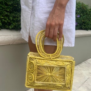May include: A yellow woven handbag with a rectangular shape and a rounded handle. The bag features intricate woven patterns and a structured design. The bag is held by a person wearing a white dress.