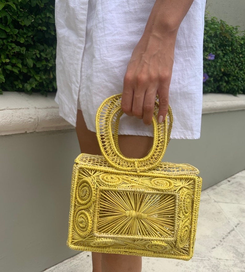 May include: A yellow woven handbag with a rectangular shape and a rounded handle. The bag features intricate woven patterns and a structured design. The bag is held by a person wearing a white dress.
