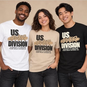 Puede incluir: Tres camisetas en blanco, beige y negro, cada una con el texto US OVER DIVISION, un gráfico de pincelada color canela y la frase HUMANITY IS GREATER THAN WHAT DIVIDES US impresa en letras negras o blancas en la parte frontal.
