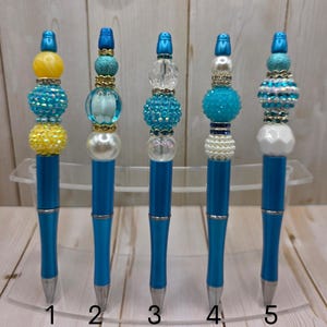 May include: Five blue pens with decorative beaded tops in various designs. The pens have silver tips and are displayed on a clear acrylic stand. The beaded tops feature a variety of colors, including yellow, turquoise, and white.