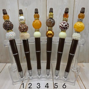 May include: Six decorative pens with brown barrels and silver accents are displayed in a clear holder. Each pen features a unique beaded design in shades of brown, gold, and white. The pens are numbered 1 through 6.