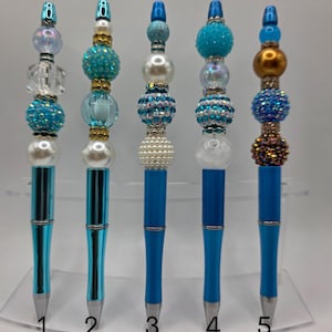 May include: Five blue and silver pens with decorative beaded tops. Each pen features a unique combination of beads in shades of blue, white, and gold. The pens are displayed on a clear, angled stand.