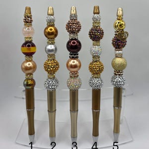 Könnte beinhalten: Fünf dekorative Stifte mit Perlenmustern in verschiedenen Farben, darunter Gold, Silber, Braun und Rosa. Jeder Stift hat eine andere Perlenanordnung und einen goldfarbenen Stiftschaft. Die Stifte sind von 1 bis 5 nummeriert.