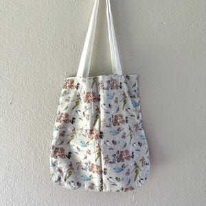 May include: A white tote bag with white straps, hanging against a light gray wall. The bag features a repeating pattern of cartoon characters in various colors on a light gray background. The bag is made of fabric.