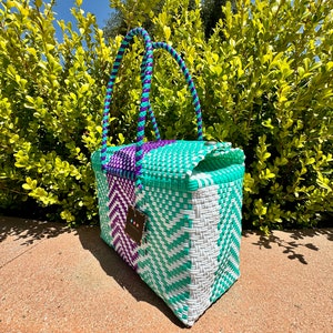 May include: A woven tote bag with purple and turquoise handles and a white and turquoise woven body. The bag is sitting on a concrete surface.