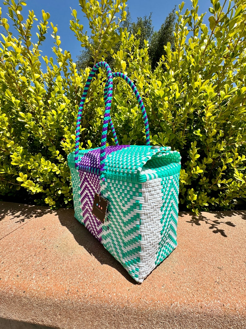 May include: A woven tote bag with purple and turquoise handles and a white and turquoise woven body. The bag is sitting on a concrete surface.