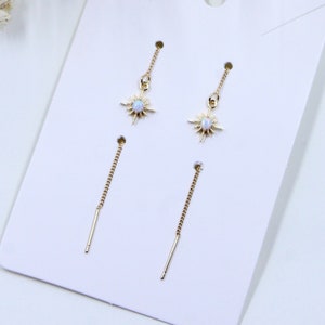 Puede incluir: Un par de pendientes de oro con un dije en forma de estrella y una piedra de ópalo blanco en el centro. Los pendientes se muestran en una tarjeta blanca.