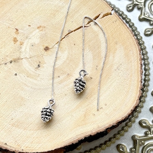May include: Silver threader earrings with miniature pinecone pendants. The earrings are presented on a wooden slice, set against a decorative, gold-coloured plate. The pinecone charms offer a touch of nature-inspired elegance.