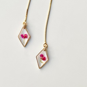 Puede incluir: Un par de pendientes de cadena dorada con un colgante en forma de diamante que presenta una flor rosa prensada.